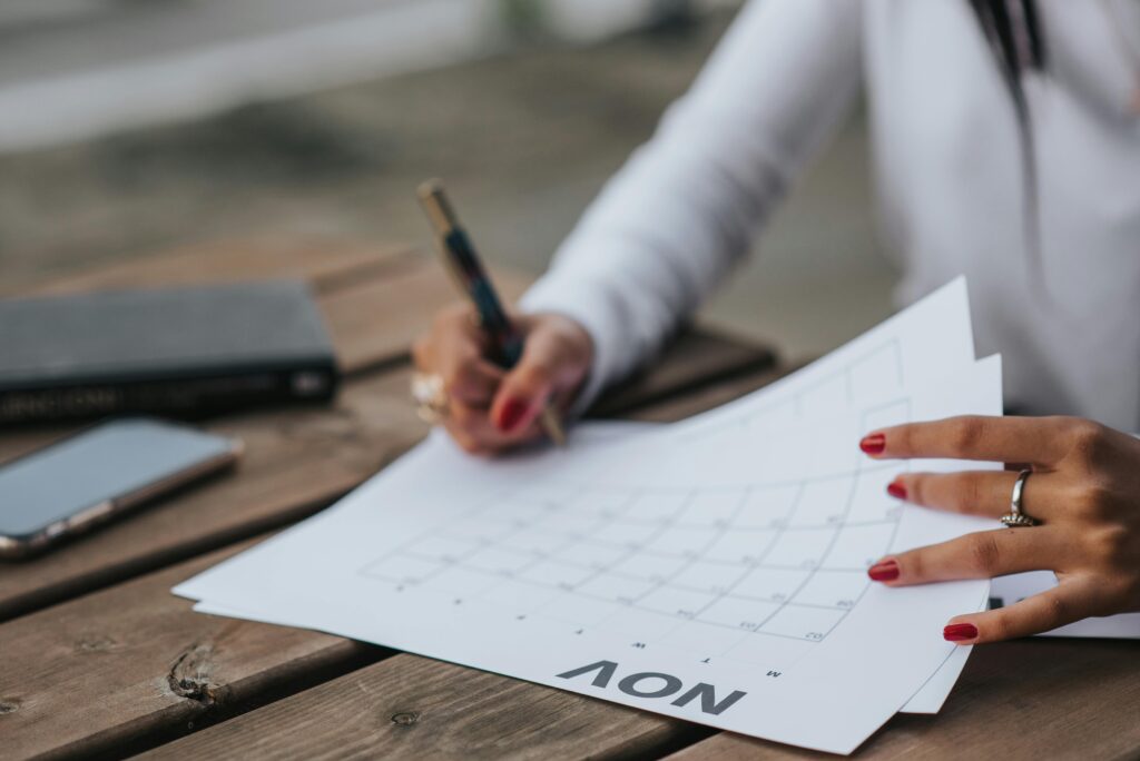 Crop faceless female in white blouse making plans for month while sitting at wooden table in park